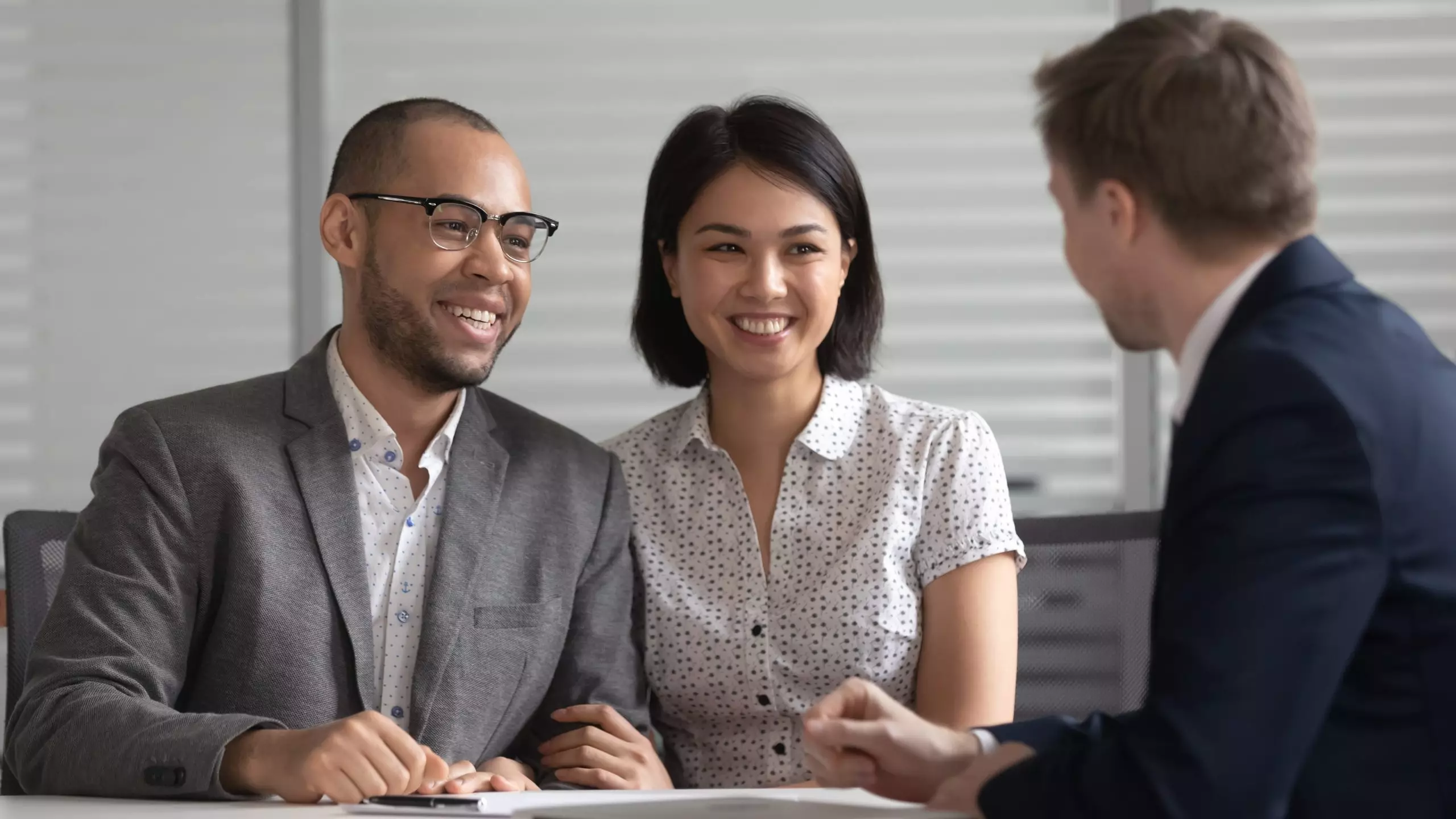 A couple, a young Black man and a young asian woman, smiling. Signing documents with a white male agent.